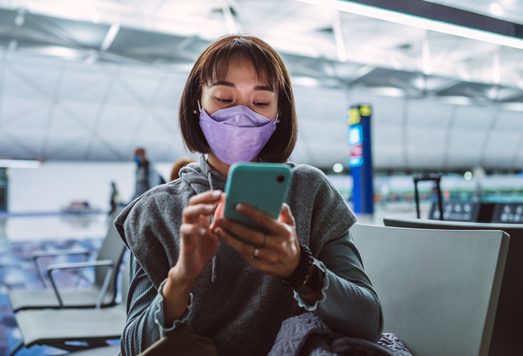 Young Asian woman in protective face mask using smartphone joyfully while waiting for night flight in airport terminal