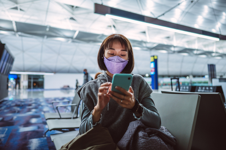 Young Asian woman in protective face mask using smartphone joyfully while waiting for night flight in airport terminal