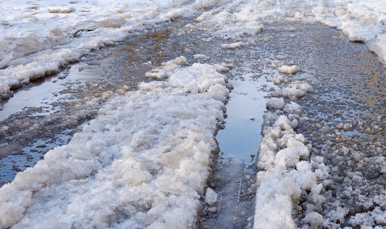 Slushy icy road surface with melting snow, deep puddles and dirty ice after winter thaw. Hazardous slippery street conditions, seasonal weather and urban winter background