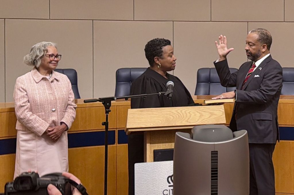 A courtroom scene with a judge administering an oath to a witness at the podium, with two other individuals present.