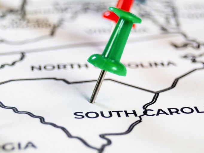 Macro shot of a green push pin on a paper map marking South Carolina. Neighboring state borders and a blurry red pin in the background symbolize East Coast travel planning and regional logistics.
