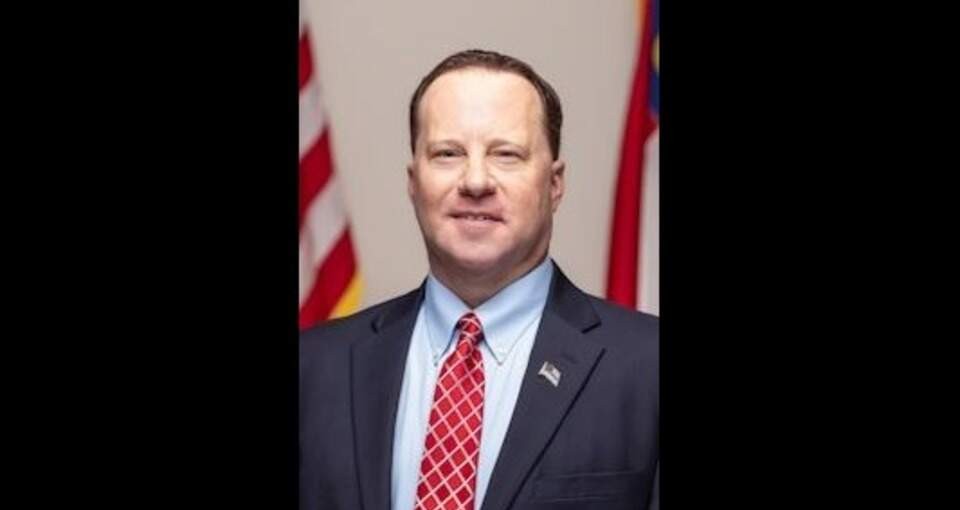 A middle-aged man in a suit and red tie standing in front of an American flag.