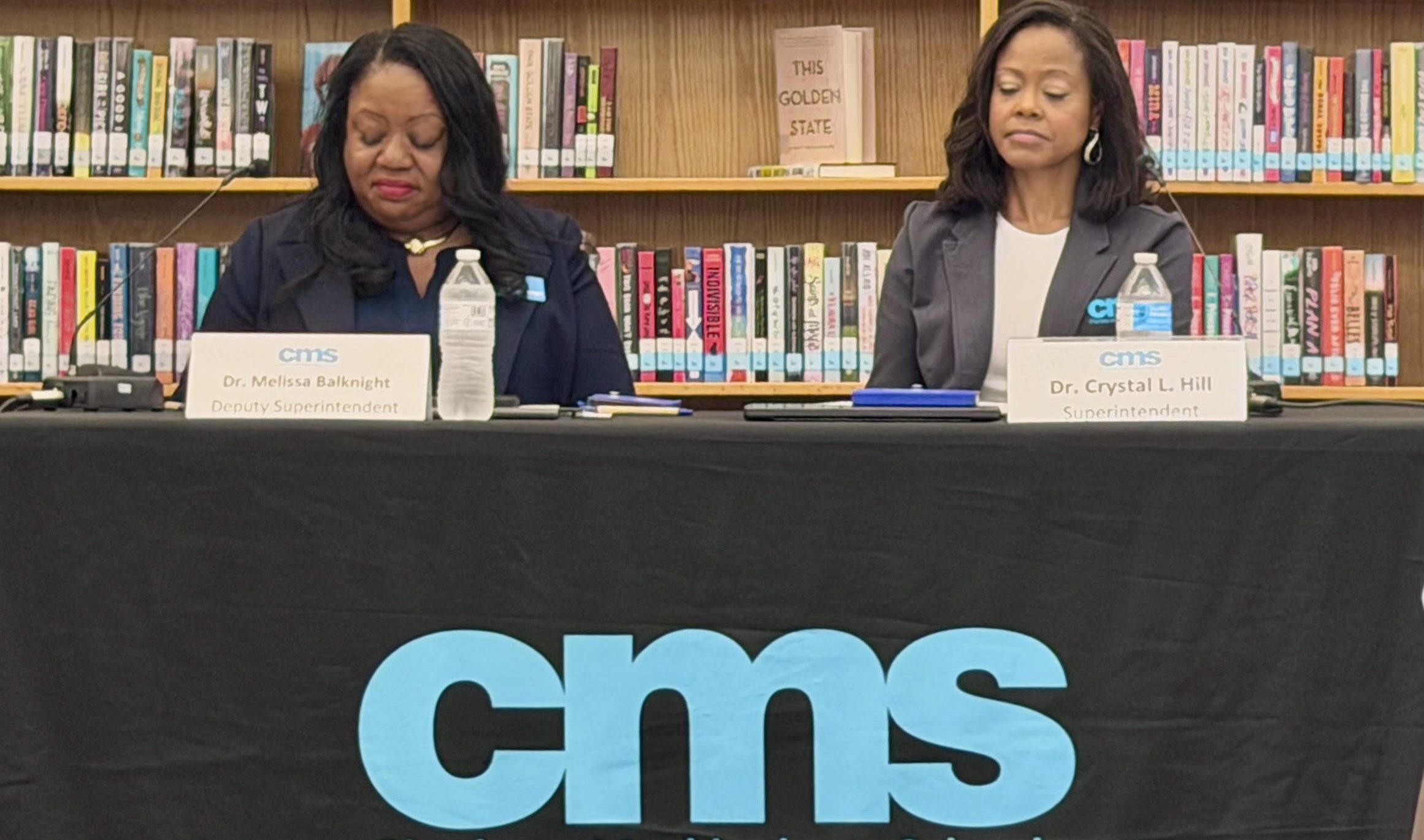 Two women seated at a table in front of bookshelves, with a "Charlotte-Mecklenburg Schools" logo displayed on the table.